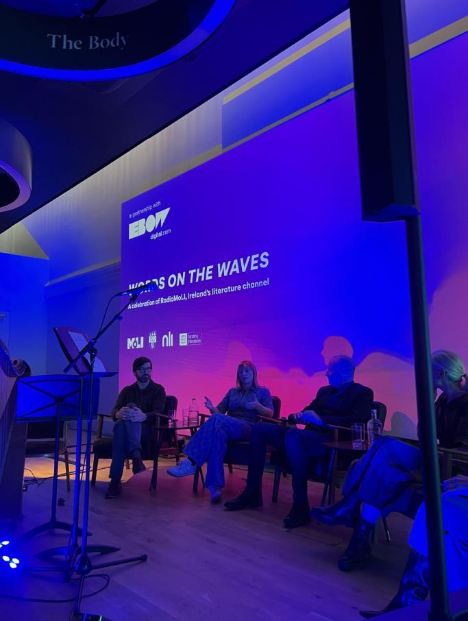 Three people sitting on chairs in front of a large lit-up screen that says ‘Words on the waves’ with a microphone stand in the foreground.
